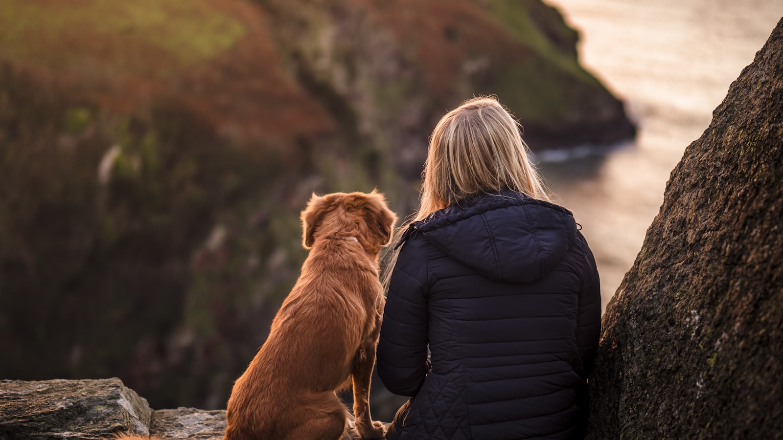 woman in black jacket sitting beside brown dog on rock near body of water during daytime